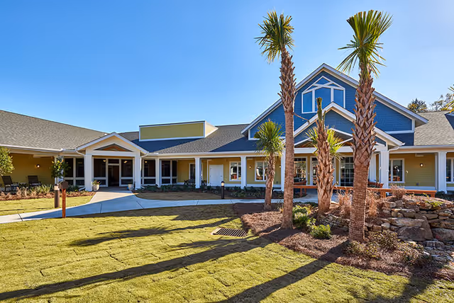 Exterior view of a single-story building with a blue and yellow facade, a covered entrance, and a landscaped front yard featuring palm trees and a rock garden under a clear blue sky.
