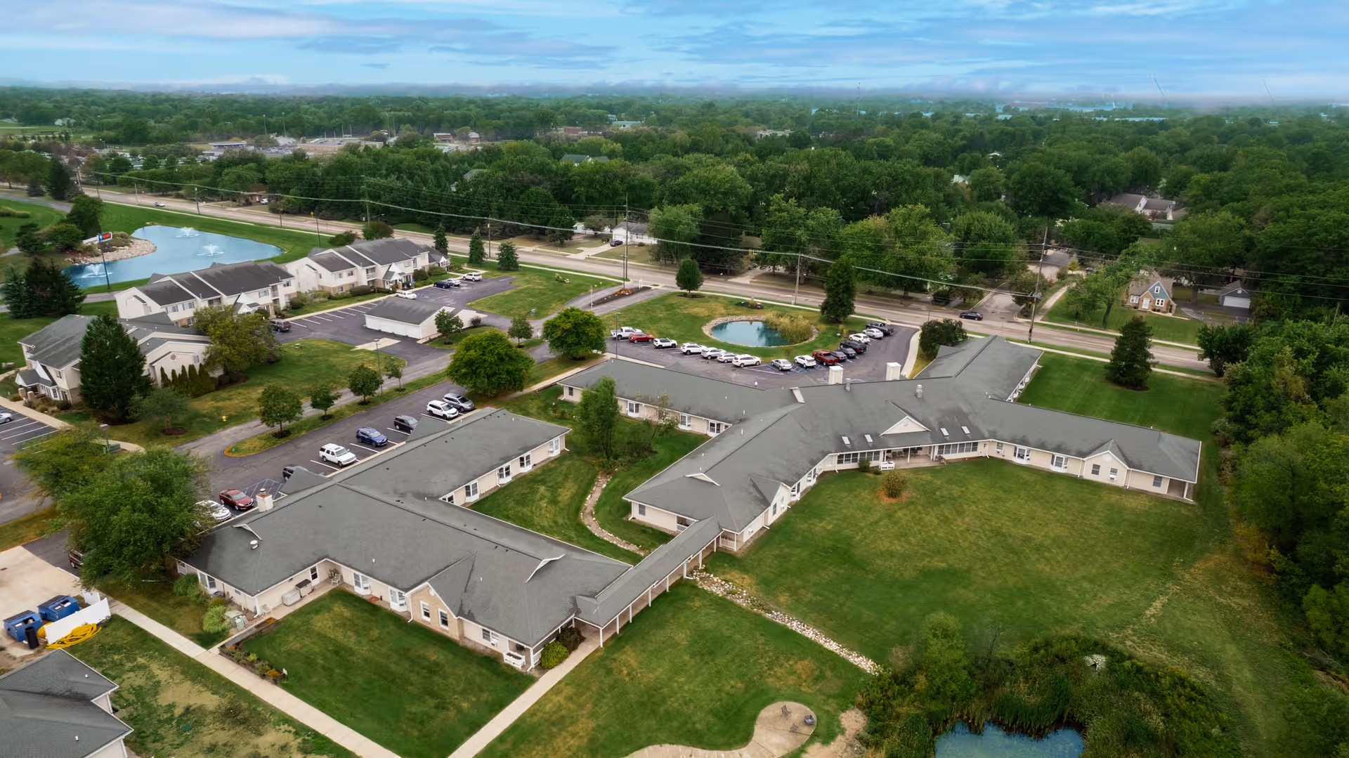 Aerial view of Addington Place of Lakeside Vista senior living facility showing multiple connected buildings with gray roofs, surrounding green lawns, parking lots with cars, small ponds, and trees under a partly cloudy sky.