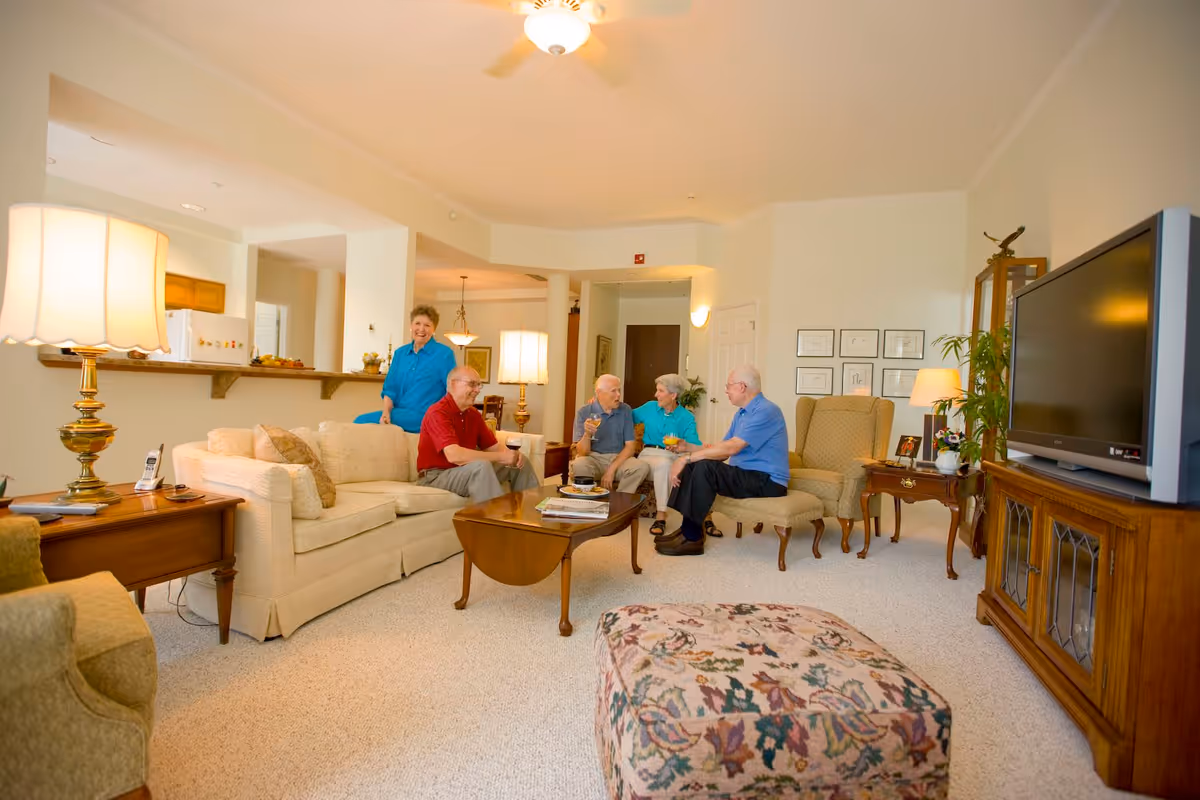A bright and spacious living room with beige carpet and cream-colored walls. The room features a beige sofa, a wooden coffee table with magazines, a floral-patterned ottoman, and a large flat-screen TV on a wooden cabinet. Four elderly people are seated and engaged in conversation, while one woman stands behind the sofa smiling. The room is warmly lit with table lamps and a ceiling fan light. The kitchen is visible in the background.