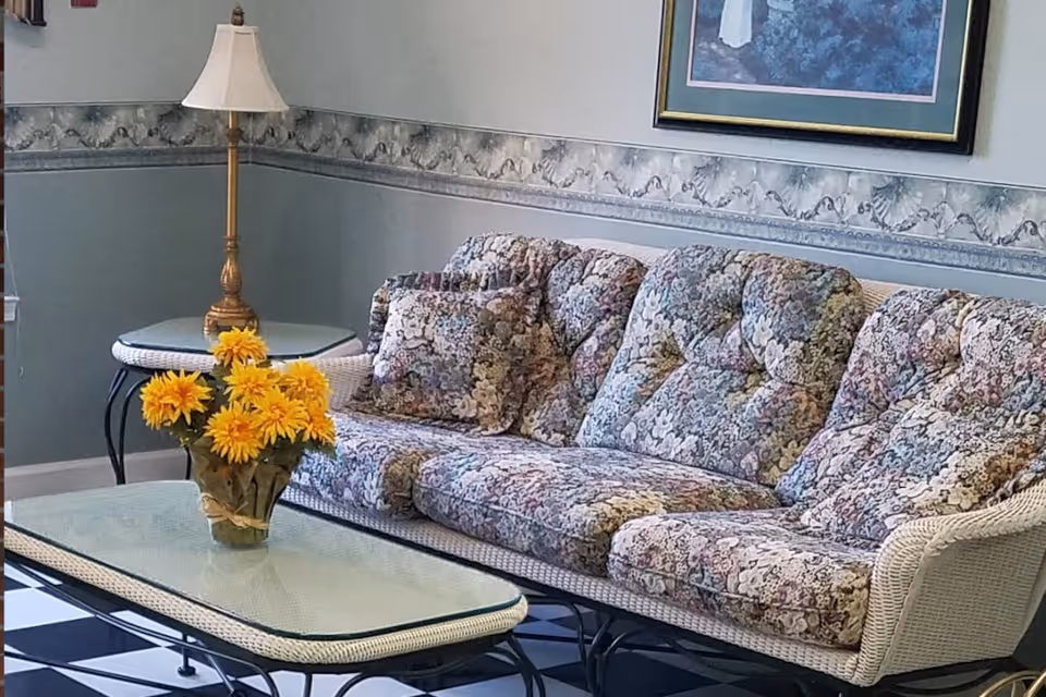 A living room area with a floral-patterned cushioned wicker sofa and matching pillows. In front of the sofa is a glass-top coffee table with a vase of bright yellow flowers. To the left, there is a side table with a lamp. The walls are decorated with a floral wallpaper border and a framed picture.