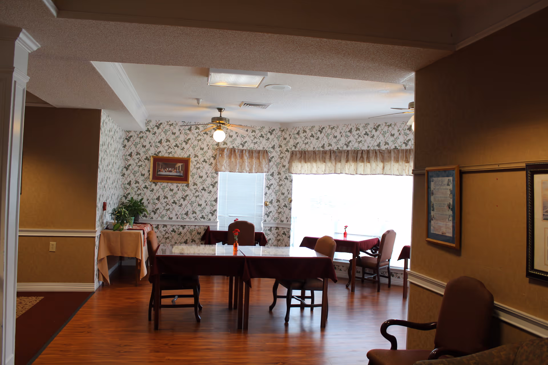 Interior dining area with tables and chairs draped in burgundy tablecloths, floral wallpaper, and ceiling fans.