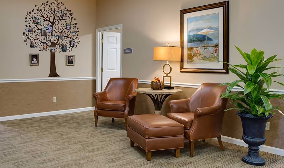 A cozy seating area in a senior living facility with two brown leather armchairs and a matching ottoman. Between the chairs is a round side table with a decorative lamp and a small pumpkin decoration. On the wall behind the chairs is a framed painting of an outdoor scene with a table and umbrella. To the left, there is a wall decal of a family tree with photos, and an open door leading to another room. A large green potted plant is placed on the right side of the seating area.