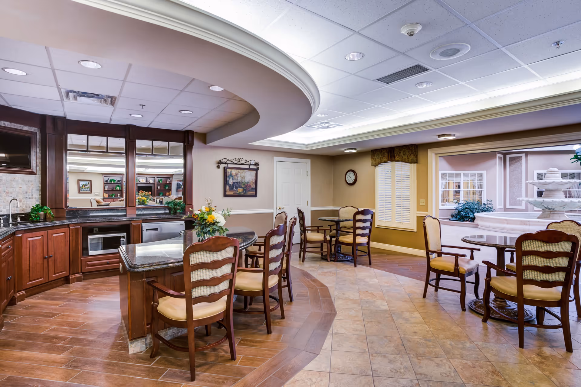 A cozy dining area in a senior living facility featuring a curved granite countertop with wooden chairs, a small kitchenette with a microwave and sink, several round tables with cushioned wooden chairs, a wall clock, a framed painting, and a large window showing an indoor courtyard with a decorative fountain.