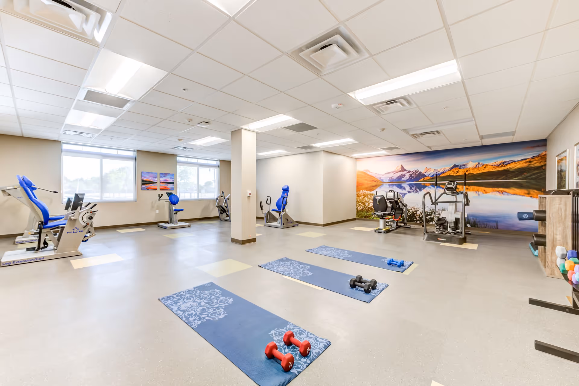 A spacious fitness room with exercise equipment including stationary bikes, weight machines, and yoga mats with dumbbells on the floor. The room has large windows letting in natural light and a large scenic mural of mountains and a lake on one wall.