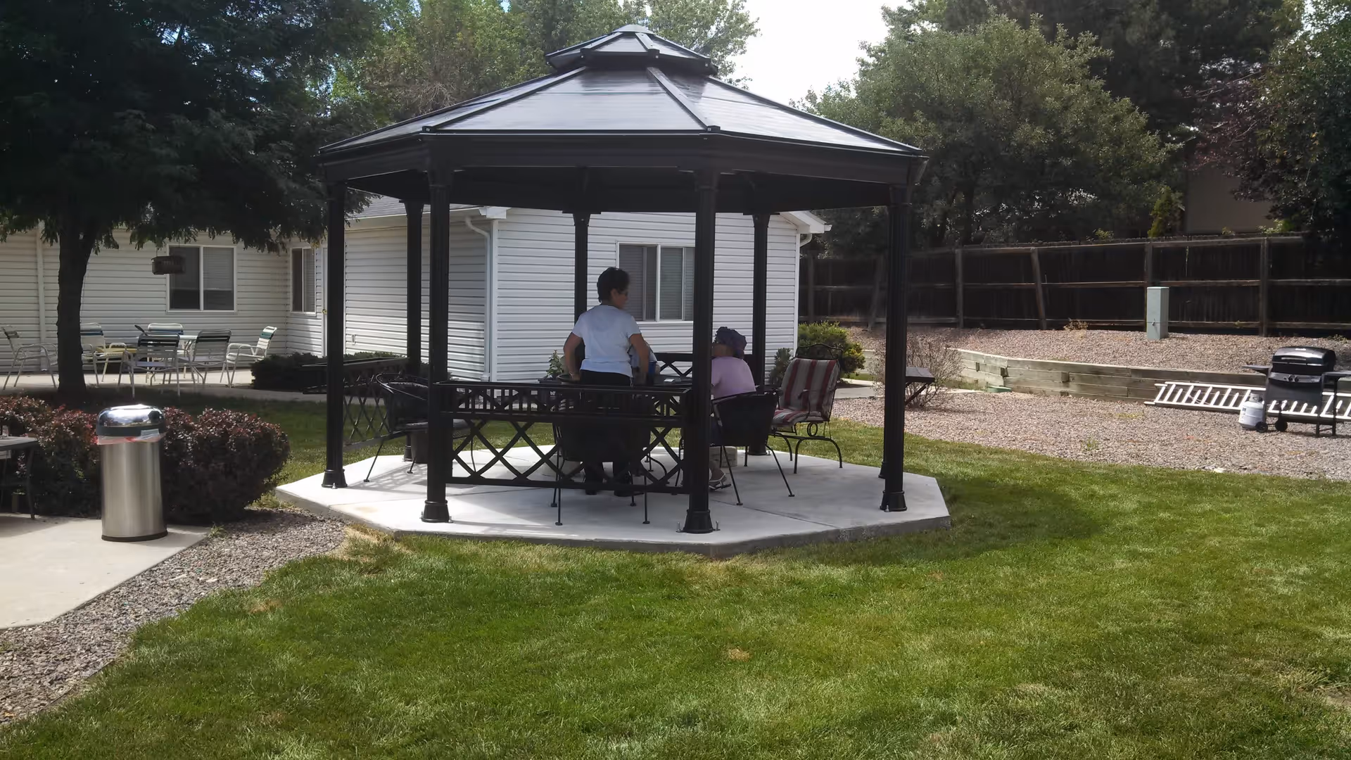Two people sitting and talking under a black metal gazebo in a grassy outdoor area with trees, bushes, and a white building in the background. There are outdoor chairs and tables nearby, a trash can, and a barbecue grill with a ladder on the gravel area.