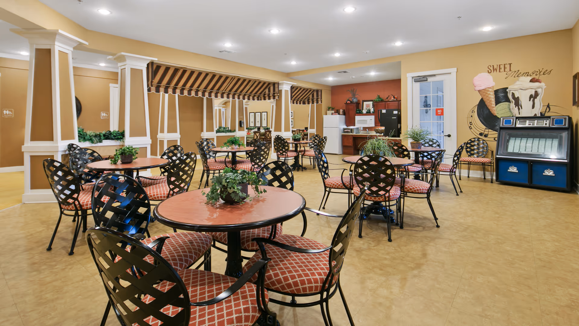 A bright and spacious dining area with multiple round tables and black metal chairs with red patterned cushions. Each table has a small green plant centerpiece. The walls are painted in warm tones with decorative columns and a large mirror. In the background, there is a kitchenette area with a refrigerator, microwave, and cabinets. On the right wall, there is a mural featuring ice cream cones, a milkshake, and a jukebox with the words 'Sweet Memories'.
