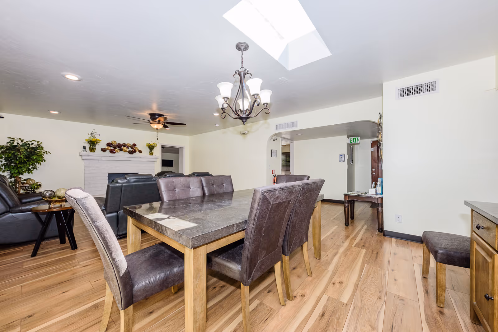 Bright communal dining area with a large wooden table and upholstered chairs opening into a living room with a fireplace and seating.