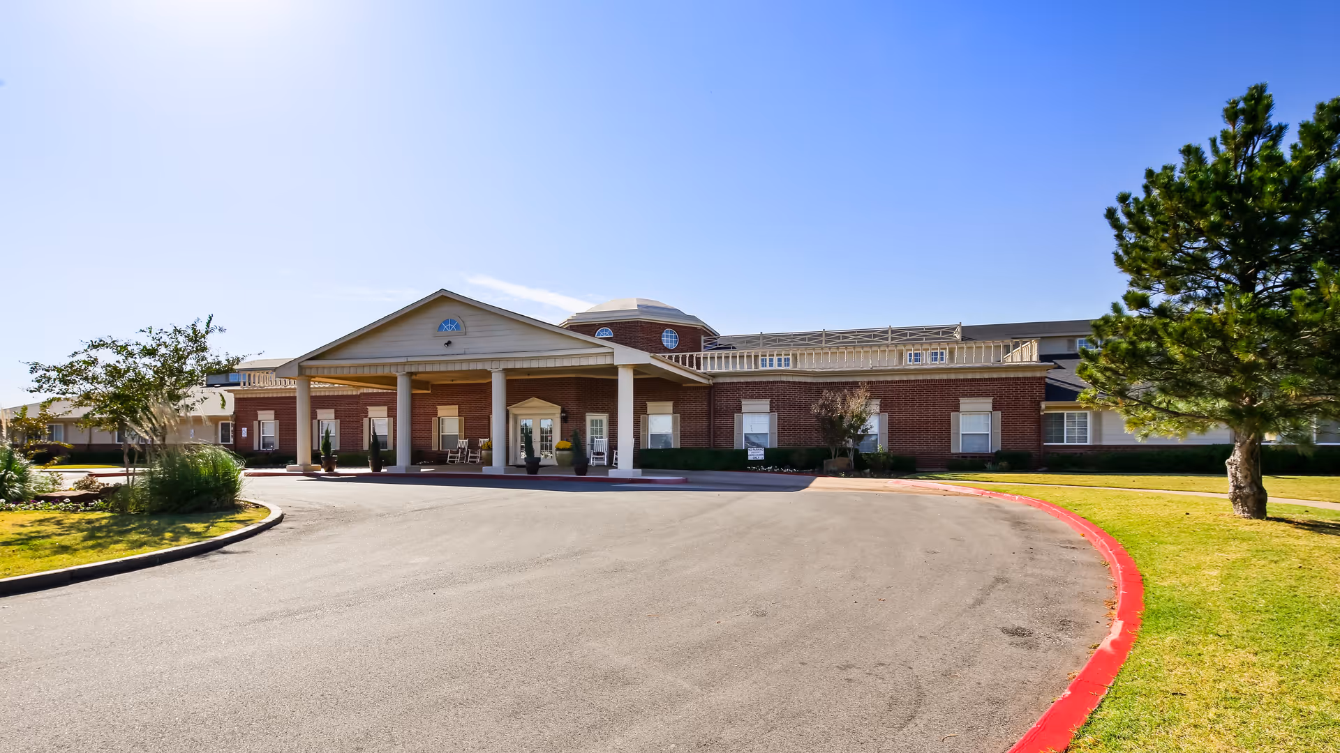 Front exterior view of a single-story brick building with a covered entrance supported by white columns, surrounded by a curved driveway and landscaped greenery under a clear blue sky.