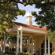Exterior view of a building with a covered entrance supported by white columns, a clock tower on the roof, and surrounded by trees with green foliage.