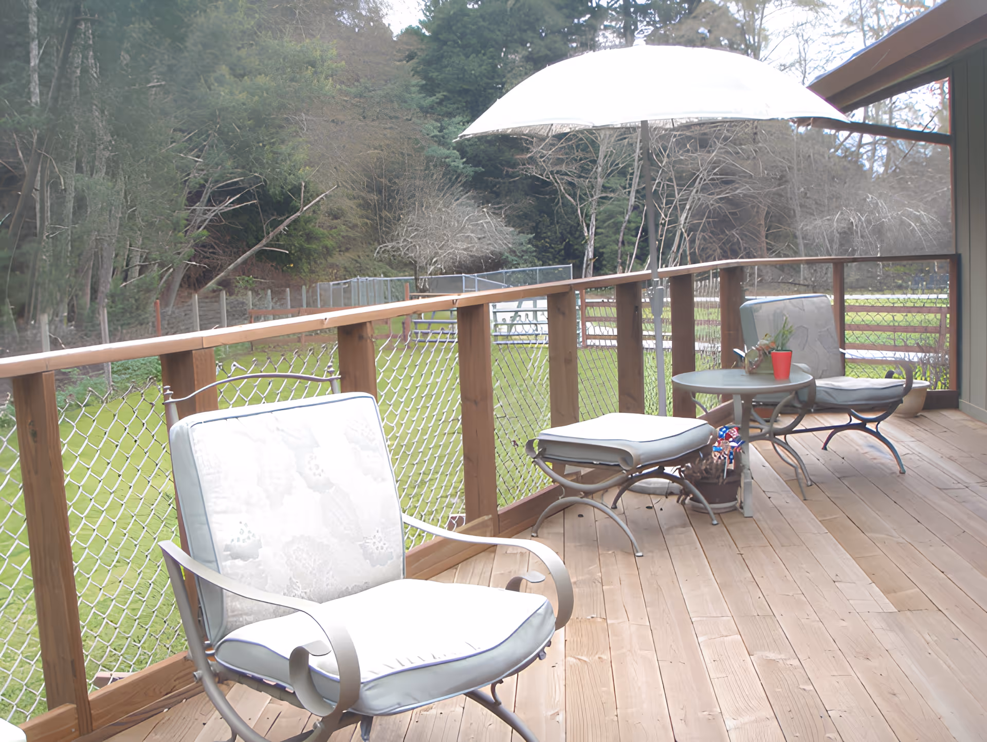 A wooden deck with two cushioned metal chairs, a small round table, and a white patio umbrella. The deck overlooks a grassy area with trees and a chain-link fence.
