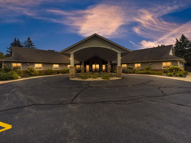 Exterior view of a single-story senior care facility building at dusk with a covered entrance supported by stone pillars, surrounded by landscaped greenery and a paved driveway.