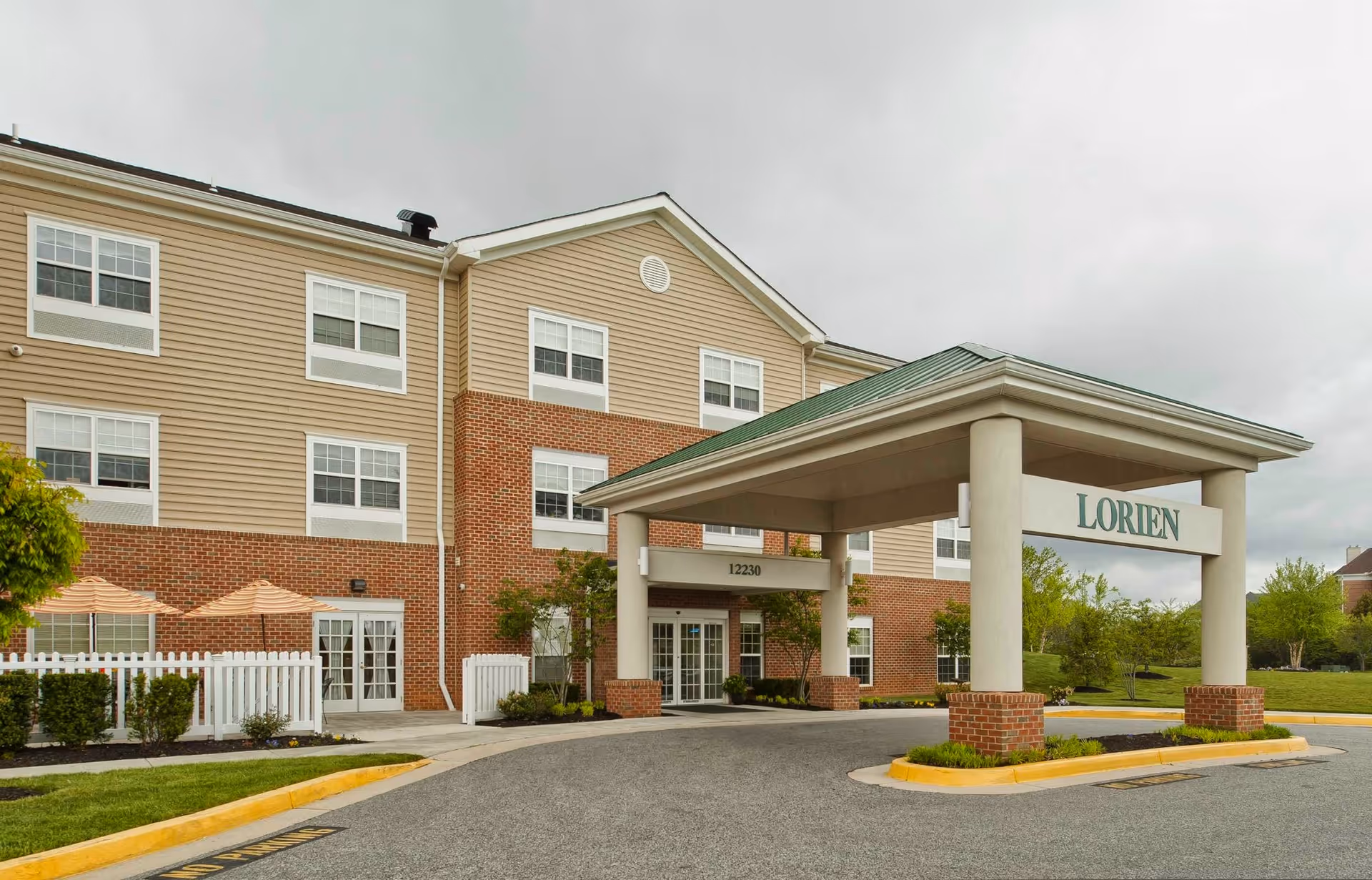 Exterior view of Lorien Mays Chapel senior living facility showing a three-story building with beige siding and red brick accents. The entrance has a covered porte-cochère with the name 'Lorien' displayed. There are windows on all floors, a white picket fence, and some greenery around the building. The sky is overcast.