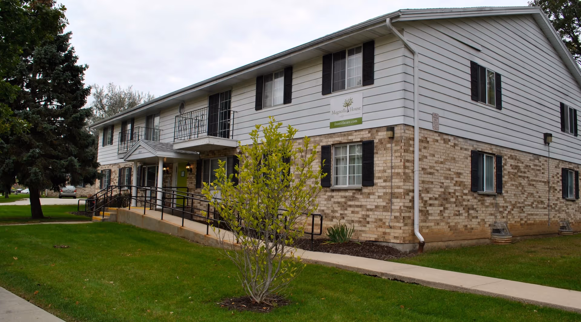 Exterior view of a two-story senior living facility building with brick and white siding, black shutters on windows, a small balcony, and a ramp leading to the entrance. There is a green lawn with a small tree and a sidewalk in front of the building. A sign on the building reads Magnolia House by Adava Care.