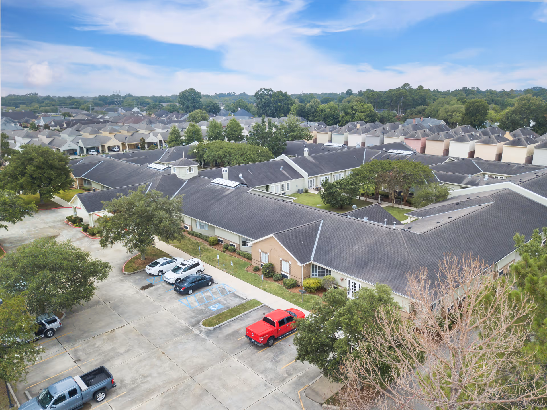Aerial view of Brookdale Lafayette senior living facility showing a large, single-story building with a dark roof, surrounded by trees and a parking lot with several cars. Residential houses and greenery are visible in the background under a partly cloudy sky.