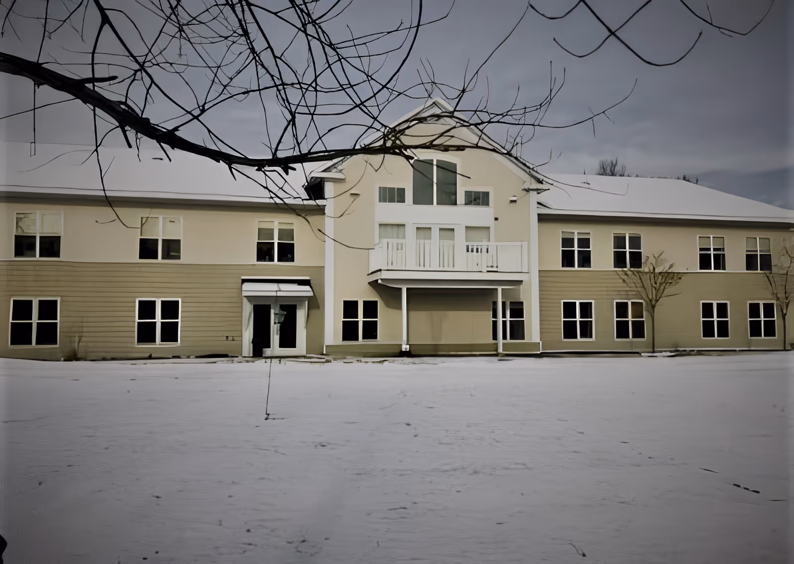 Exterior view of a two-story beige and light brown building with multiple windows and a small balcony in the center. The ground is covered with snow, and leafless tree branches are visible in the foreground.