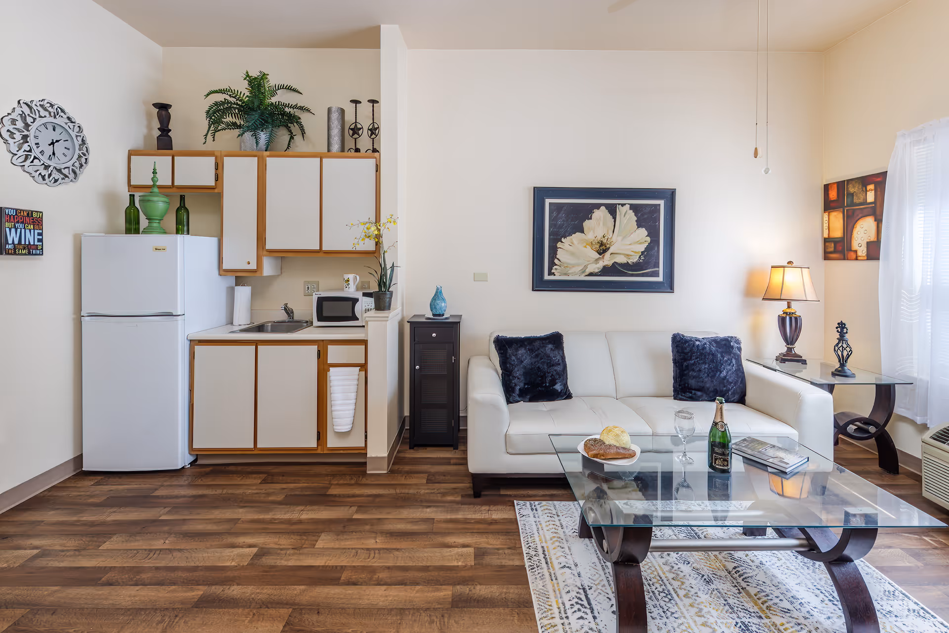 A cozy living space featuring a small kitchen area with white cabinets, a refrigerator, a microwave, and a sink. Adjacent to the kitchen is a white leather sofa with two dark blue pillows, a glass coffee table with a plate of bread, a glass of water, a bottle of champagne, and a book. The room has wooden flooring, a decorative wall clock, framed artwork, a table lamp, and a window with sheer white curtains allowing natural light to enter.