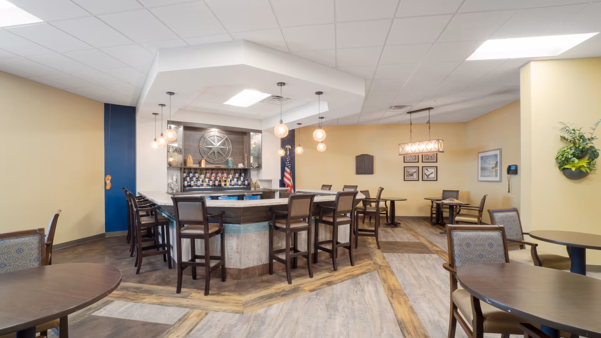 A well-lit dining area with a central bar counter surrounded by high chairs. The bar has hanging pendant lights and shelves stocked with bottles and decorative items. Several round tables with chairs are arranged around the room, which has light-colored walls, wood-patterned flooring, and framed artwork on the walls.