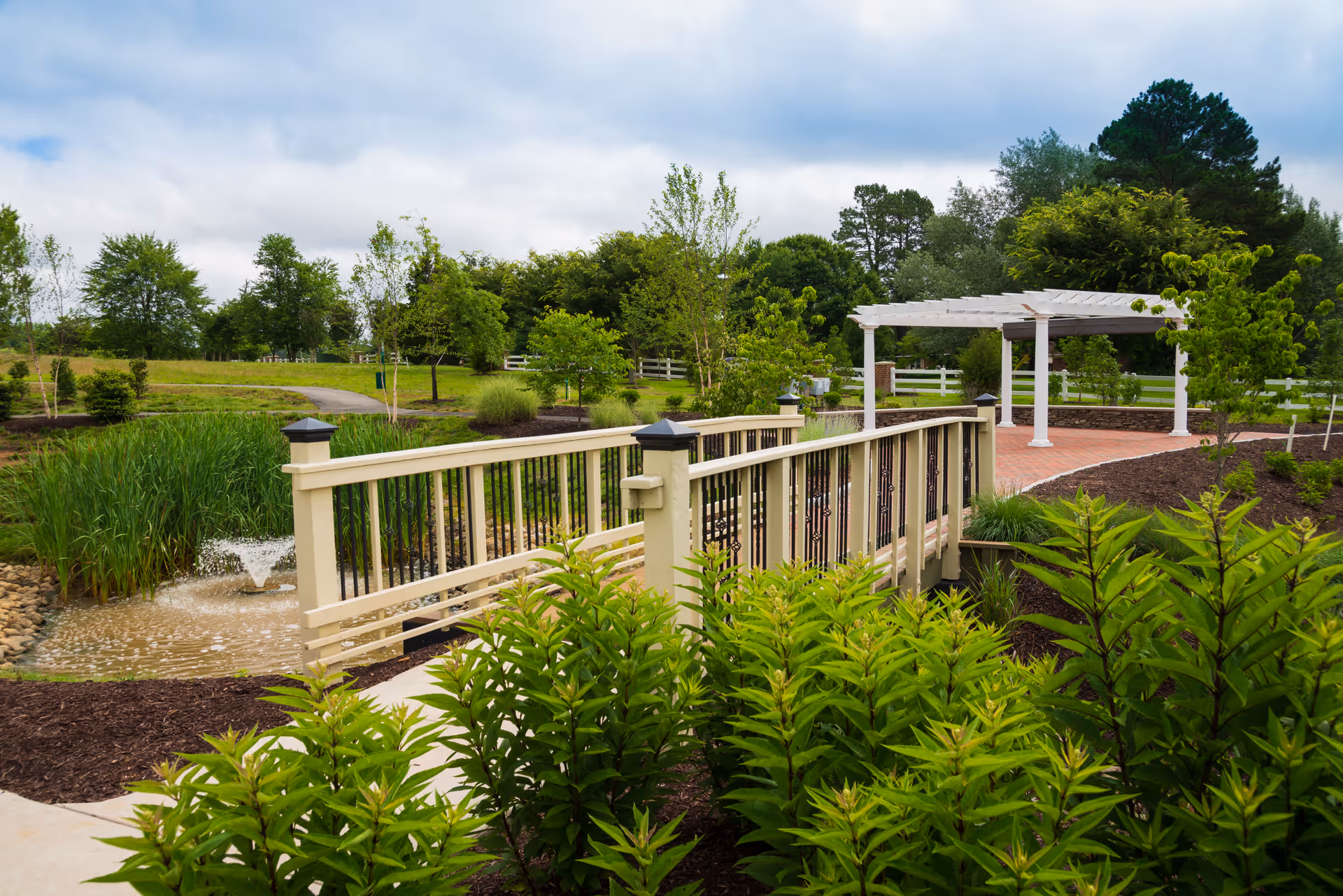 A landscaped outdoor area at Covenant Woods featuring a small beige bridge with black railings over a pond with a water fountain. Surrounding the bridge are green shrubs, trees, and a white pergola on a brick patio. The sky is partly cloudy.