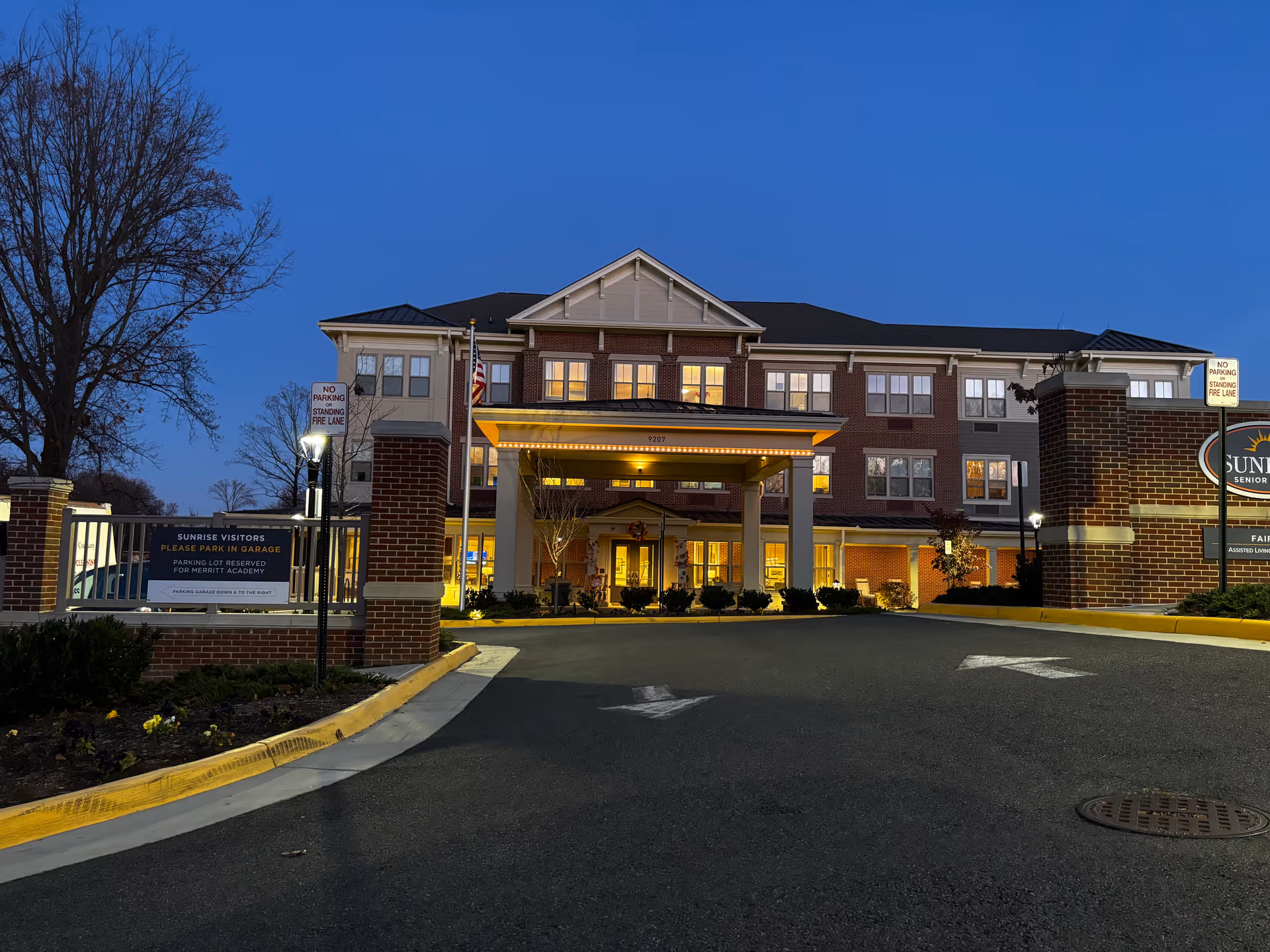 Exterior view of the Sunrise of Fairfax senior living facility at dusk, showing a three-story brick and siding building with lit windows and a covered entrance. There are signs indicating no parking or standing in the fire lane and a sign directing visitors to park in the garage. Trees and landscaping surround the driveway leading up to the entrance.