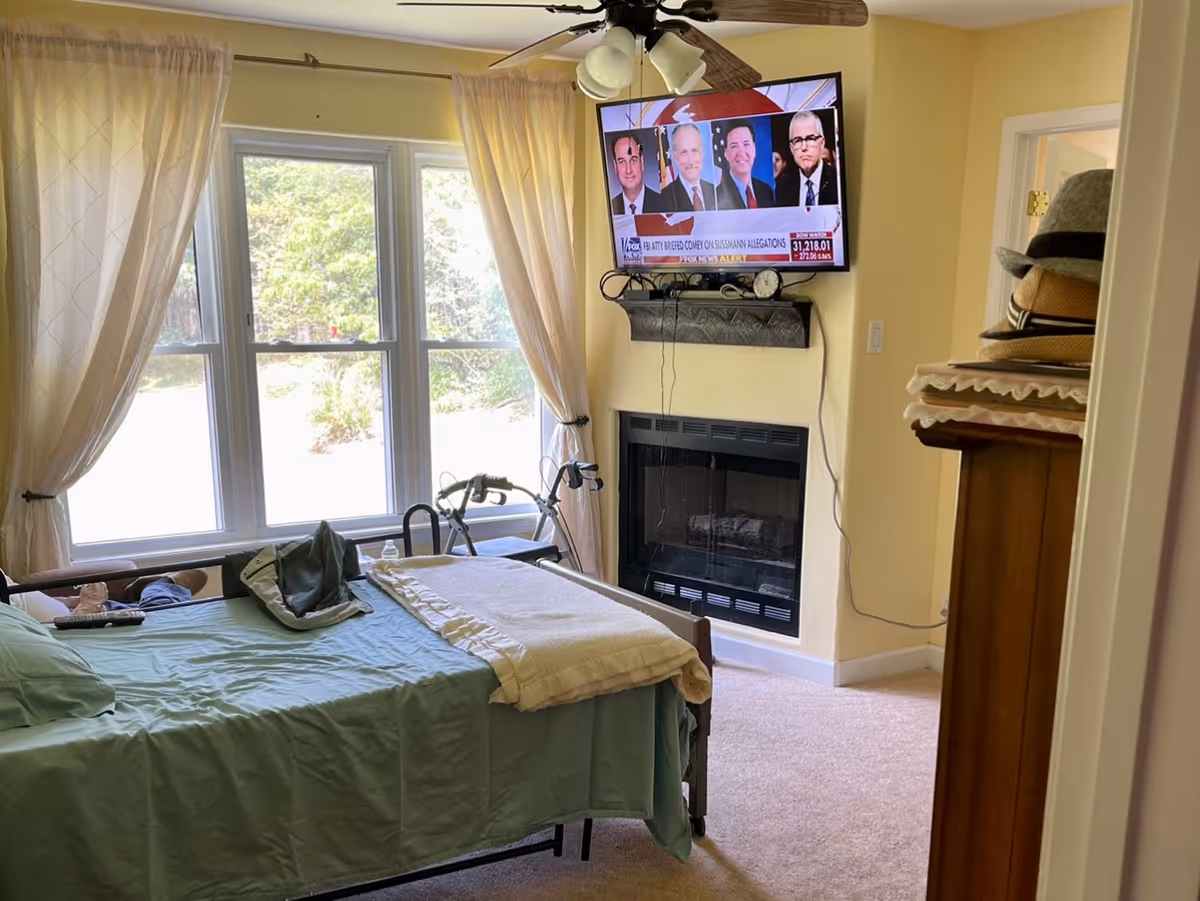 A bedroom in an assisted living facility with a bed covered in green bedding and a white blanket. There is a large window with sheer curtains letting in natural light. A walker is positioned near the window. A flat-screen TV is mounted above a fireplace, displaying a news program. A wooden dresser with hats on top is visible on the right side of the room.