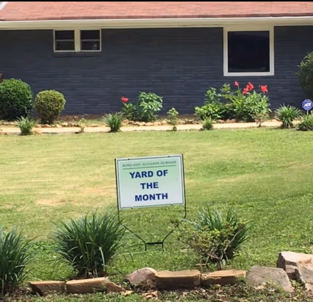 Front yard of a house with a 'YARD OF THE MONTH' sign in the grass and shrubs in front of a dark exterior wall.