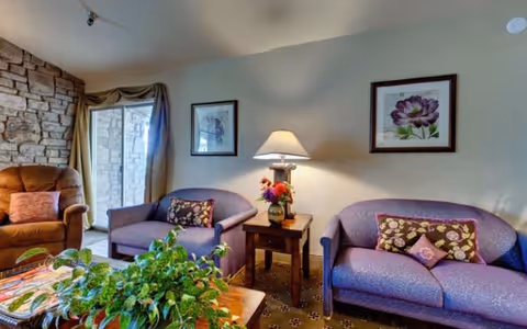 Cozy sitting area with two purple loveseats, a lamp-topped side table, a brown armchair, framed artwork, and a coffee table with plants.