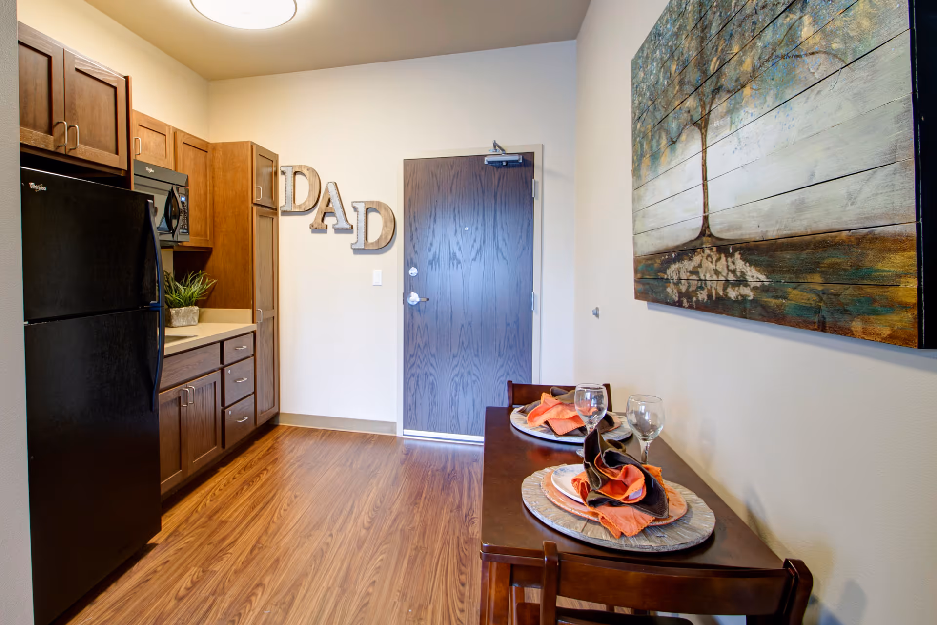 A small dining area and kitchen in a senior living facility. The kitchen features wooden cabinets, a black refrigerator, and a microwave. A wooden dining table set for two with plates, napkins, and wine glasses is positioned against the wall. Above the table hangs a large painting of a tree. The wall near the door has decorative letters spelling 'DAD'. The floor is wood laminate.