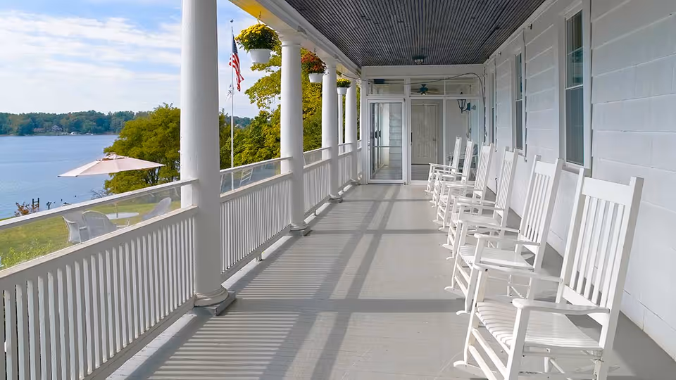 A long covered porch with white rocking chairs lined up along the wall, overlooking a scenic lake with trees and an American flag visible in the background. There are hanging flower pots attached to the porch ceiling and a patio umbrella with chairs on the lawn below.