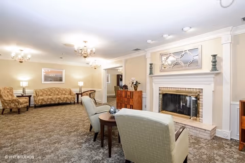 A cozy living room area with beige walls and carpeted floor. The room features two light green armchairs with a small round wooden table between them, a fireplace with decorative items on the mantel, a wooden cabinet, and a seating area with a patterned sofa and armchair. The room is softly lit by ceiling chandeliers and table lamps.
