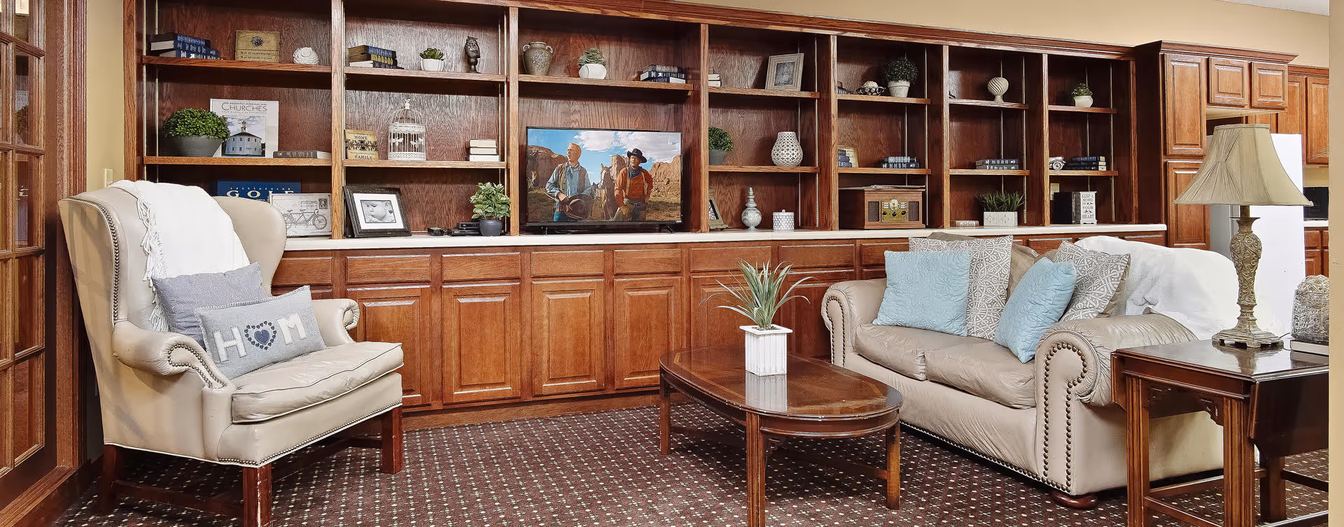 A cozy living room area with a beige leather sofa adorned with blue and patterned pillows, a beige armchair with a white throw and a decorative pillow, a wooden coffee table with a small potted plant, and a wooden side table with a lamp. Behind the seating is a large wooden built-in bookshelf with various decorative items and a television displaying a western movie scene.