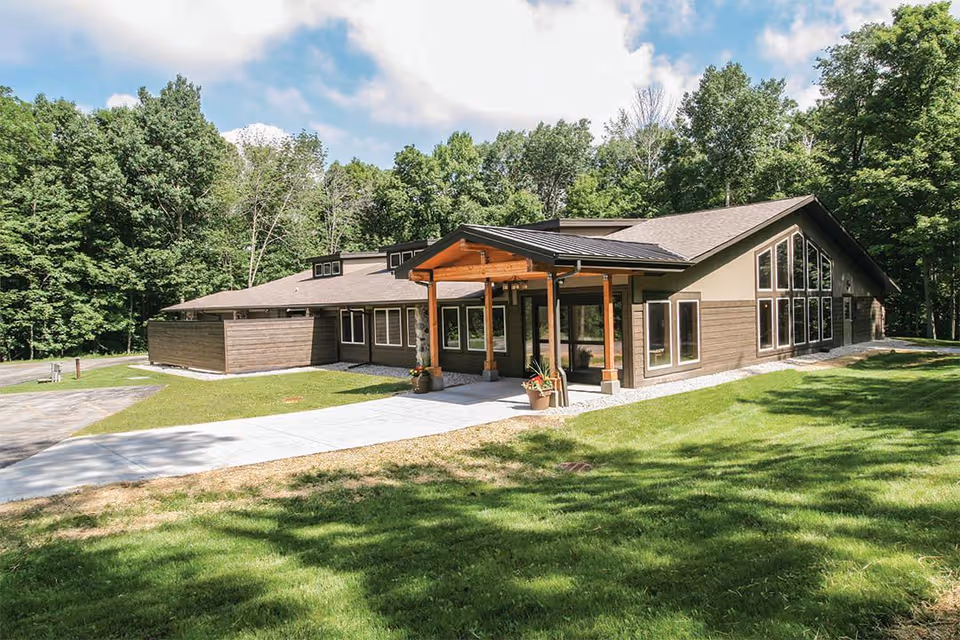 Exterior view of a single-story building with a modern design, featuring large windows and a covered entrance supported by wooden pillars. The building is surrounded by green grass and trees under a partly cloudy sky.