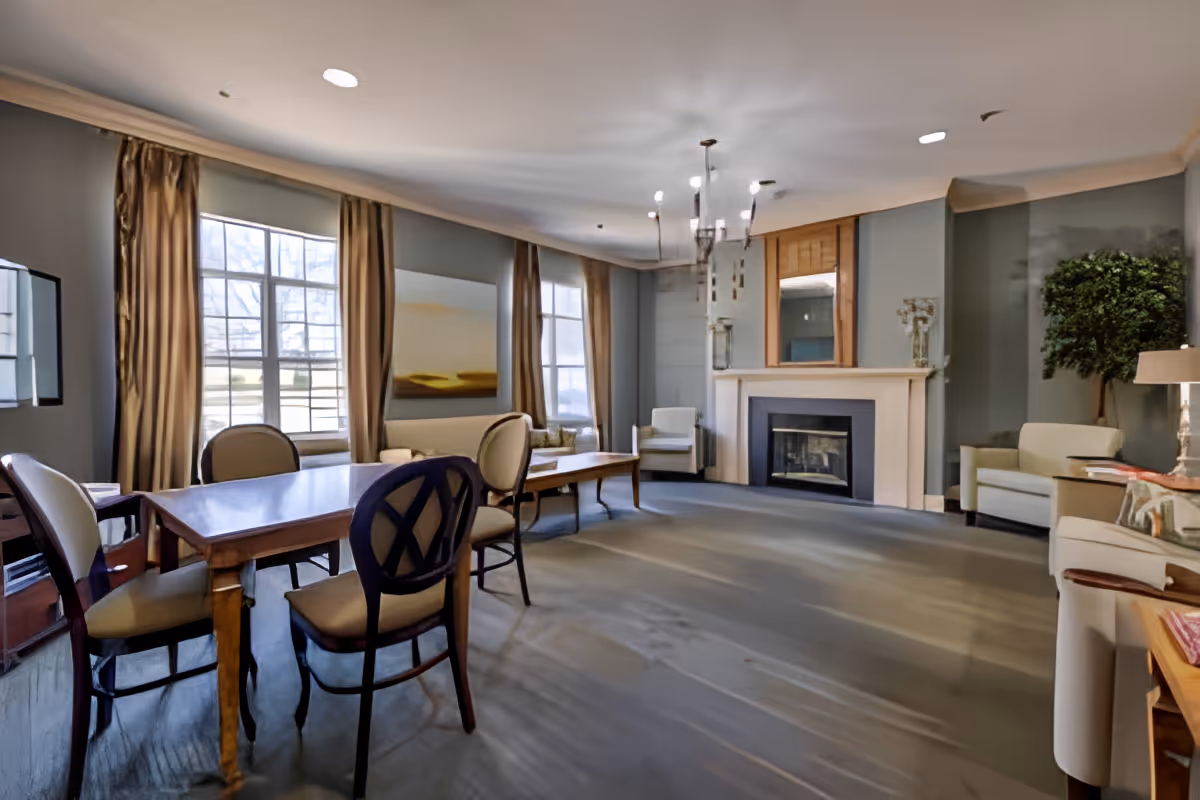 A spacious living room with large windows covered by beige curtains allowing natural light to fill the room. The room features a wooden table with four chairs, a fireplace with a mantel, two armchairs, a sofa, a coffee table, a floor lamp, and a potted plant in the corner. The walls are painted gray and there is a modern chandelier hanging from the ceiling.