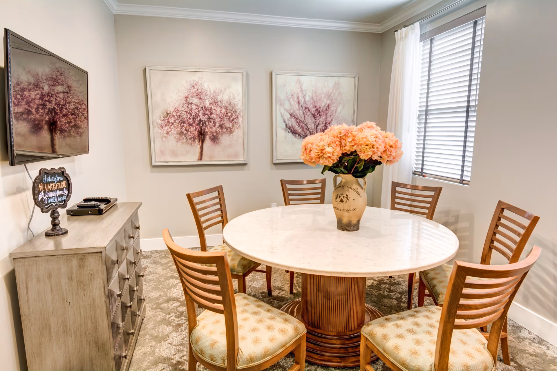 A small dining room with a round marble table surrounded by six wooden chairs with patterned cushions. A vase with pink flowers is placed in the center of the table. On the left side, there is a wooden cabinet with a small decorative sign and a flat-screen TV mounted above it. Two framed paintings of pink trees hang on the wall behind the table. A window with blinds and sheer curtains is on the right side.