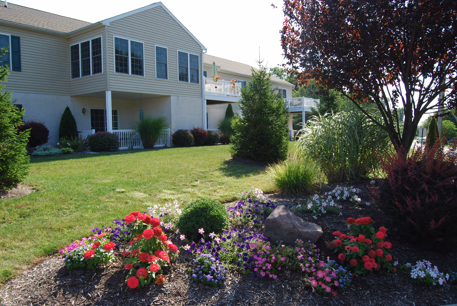 A landscaped garden area with colorful flowers, shrubs, and a small tree in front of a beige two-story building with balconies and large windows under a clear sky.