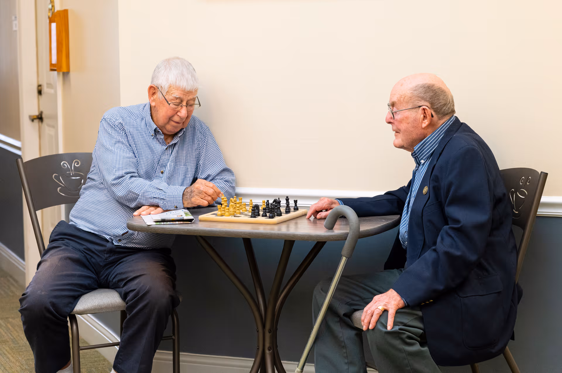 Two elderly men sitting at a small table playing chess in a well-lit indoor setting. One man is making a move while the other watches attentively. Both are dressed casually, and one has a cane resting against the table. The chairs have a decorative coffee cup design on the backrests.