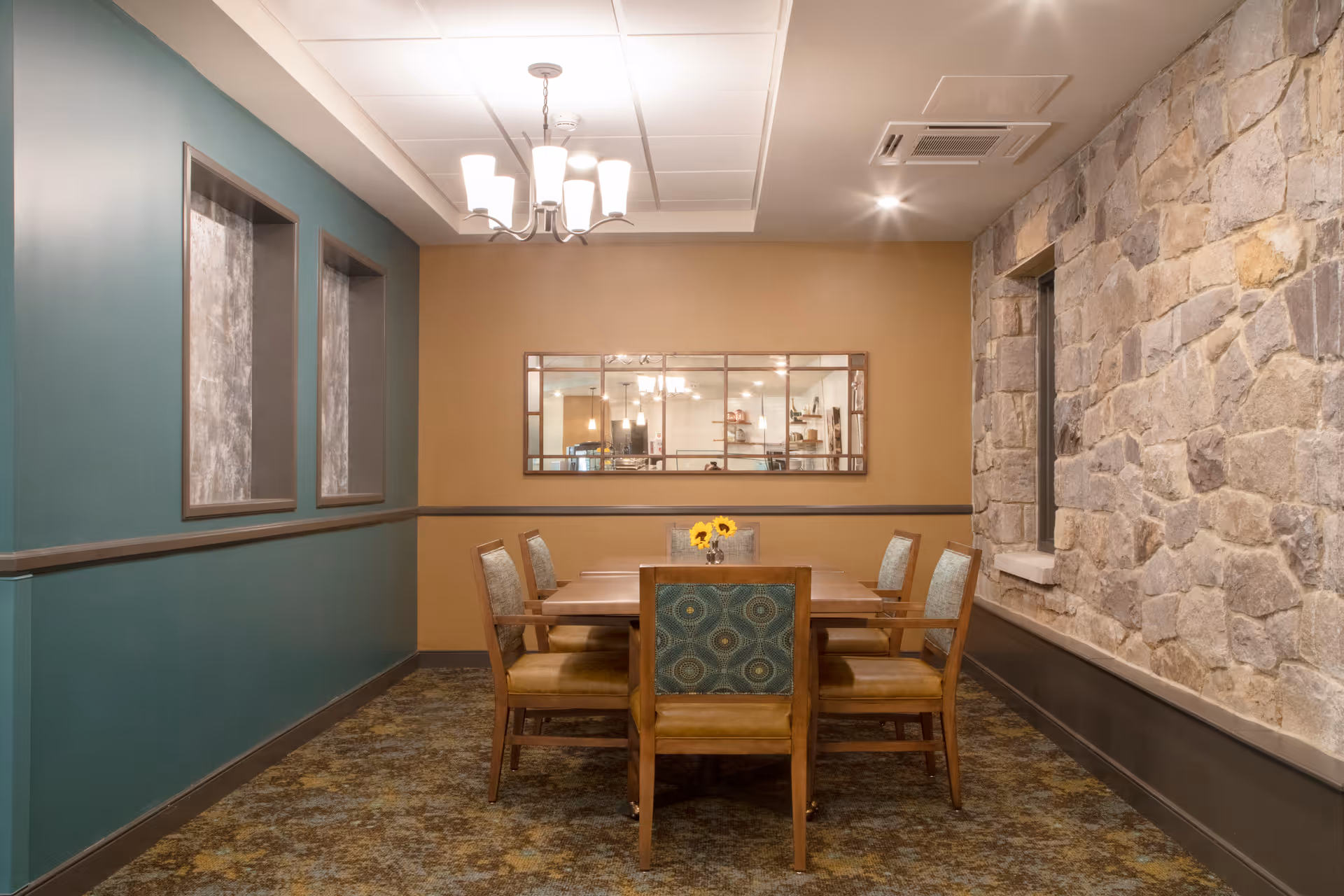A dining room with a rectangular wooden table surrounded by six chairs with patterned upholstery. The room features a stone accent wall on the right, teal and beige painted walls, a chandelier hanging from the ceiling, and a large rectangular mirror on the beige wall reflecting part of the room.