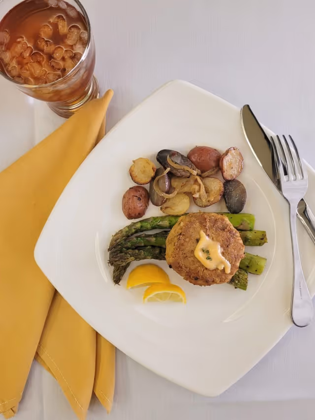 A plated meal consisting of roasted asparagus, a crab cake topped with a dollop of sauce, roasted baby potatoes with onions, and two lemon wedges on a white square plate. A glass of iced tea and a yellow cloth napkin are placed beside the plate, with a fork and knife resting on the right side of the plate.