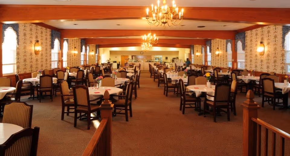 Large formal dining room with rows of round tables set with white tablecloths, wooden chairs, chandeliers, and warm wood accents.