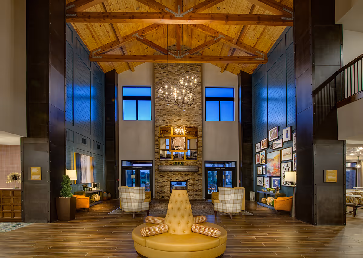 Spacious two-story senior living lobby with a vaulted wooden ceiling, central circular seating, fireplace and multiple cozy seating areas.