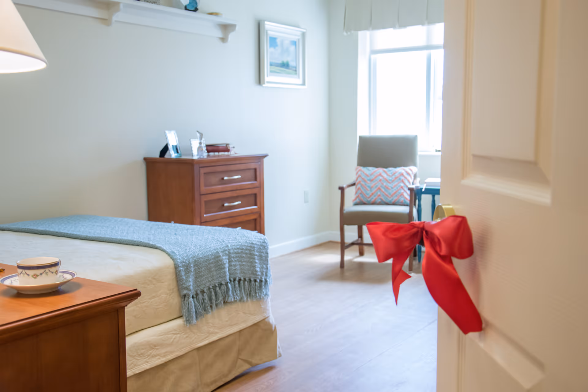A cozy senior living bedroom with a neatly made bed covered with a light blue throw blanket, a wooden nightstand with a teacup and saucer, a wooden dresser with framed photos and decorative items, and a comfortable chair with a patterned pillow near a window letting in natural light. A red ribbon bow is tied to the door handle.