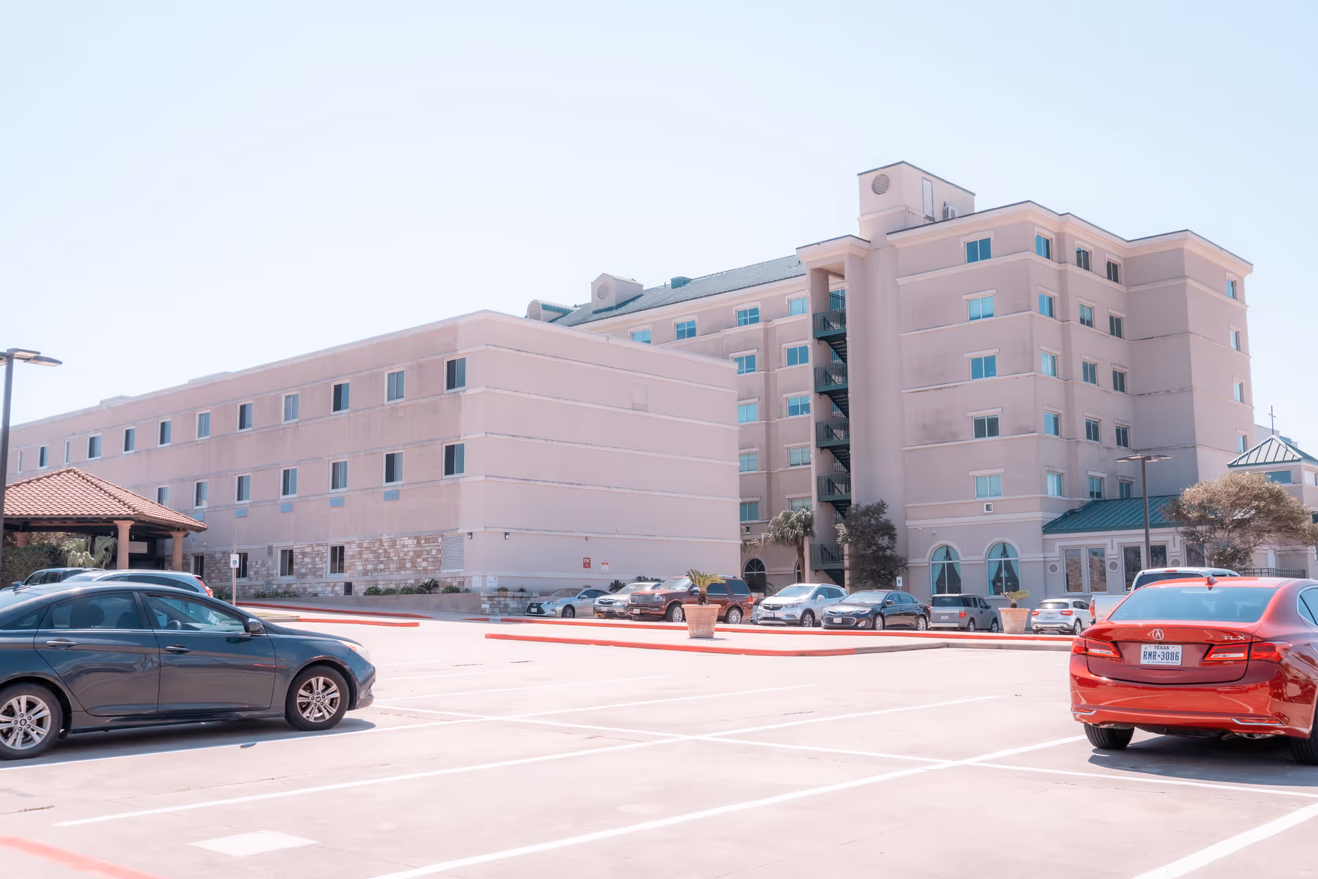 Front exterior of a multi-story senior living building with a mostly empty parking lot and several parked cars.