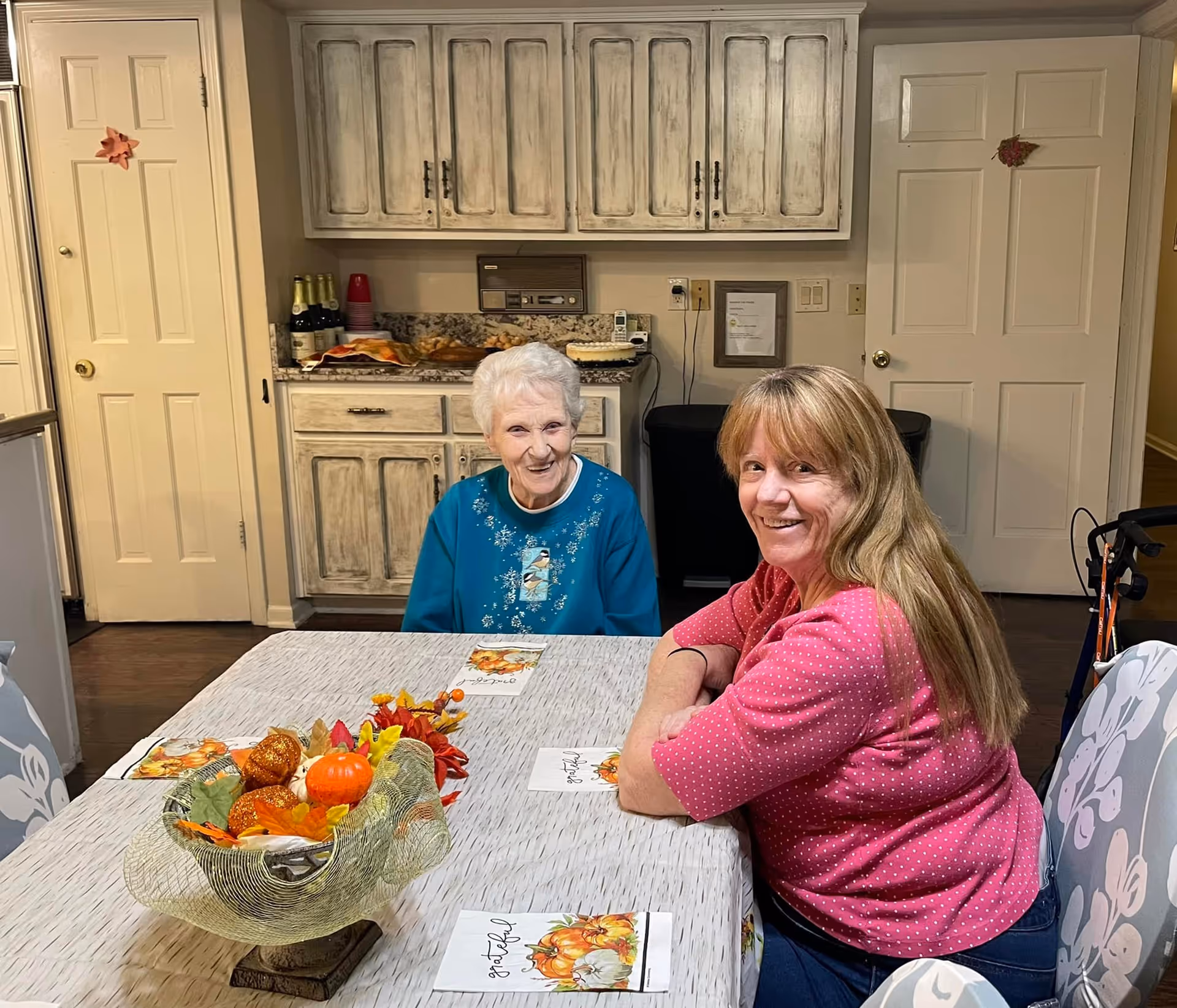 Two women sitting at a dining table in a kitchen area. One elderly woman with white hair is wearing a blue sweater and smiling, while a younger woman with long blonde hair in a pink polka dot shirt is also smiling. The table is decorated with a fall-themed centerpiece and napkins. Behind them are white distressed cabinets, a countertop with bottles and cups, and two closed doors.