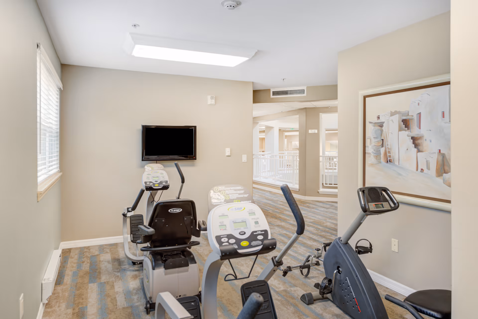 A small exercise room with two stationary recumbent bikes and one upright stationary bike. The room has beige walls, a window with blinds on the left, a wall-mounted TV, and a framed painting on the right wall. The floor is carpeted with a patterned design, and there is an open hallway visible in the background.