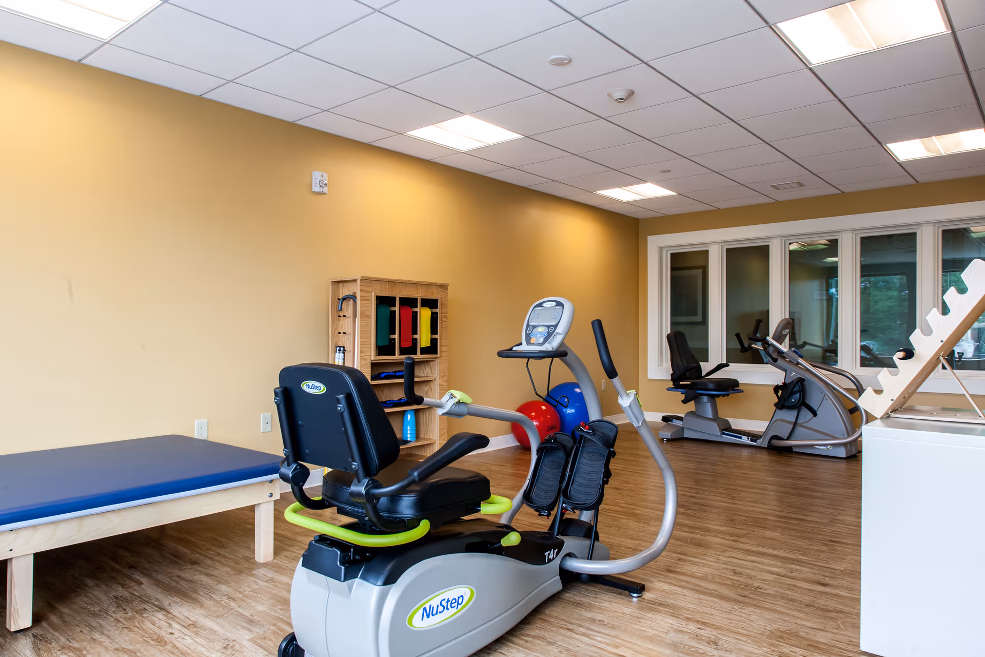 A fitness room with exercise equipment including a NuStep recumbent cross trainer, a stationary bike, exercise balls, and a padded exercise table. The room has wood flooring, beige walls, and a ceiling with recessed lighting. There is a wooden shelf with colorful exercise bands and a large window with multiple panes.