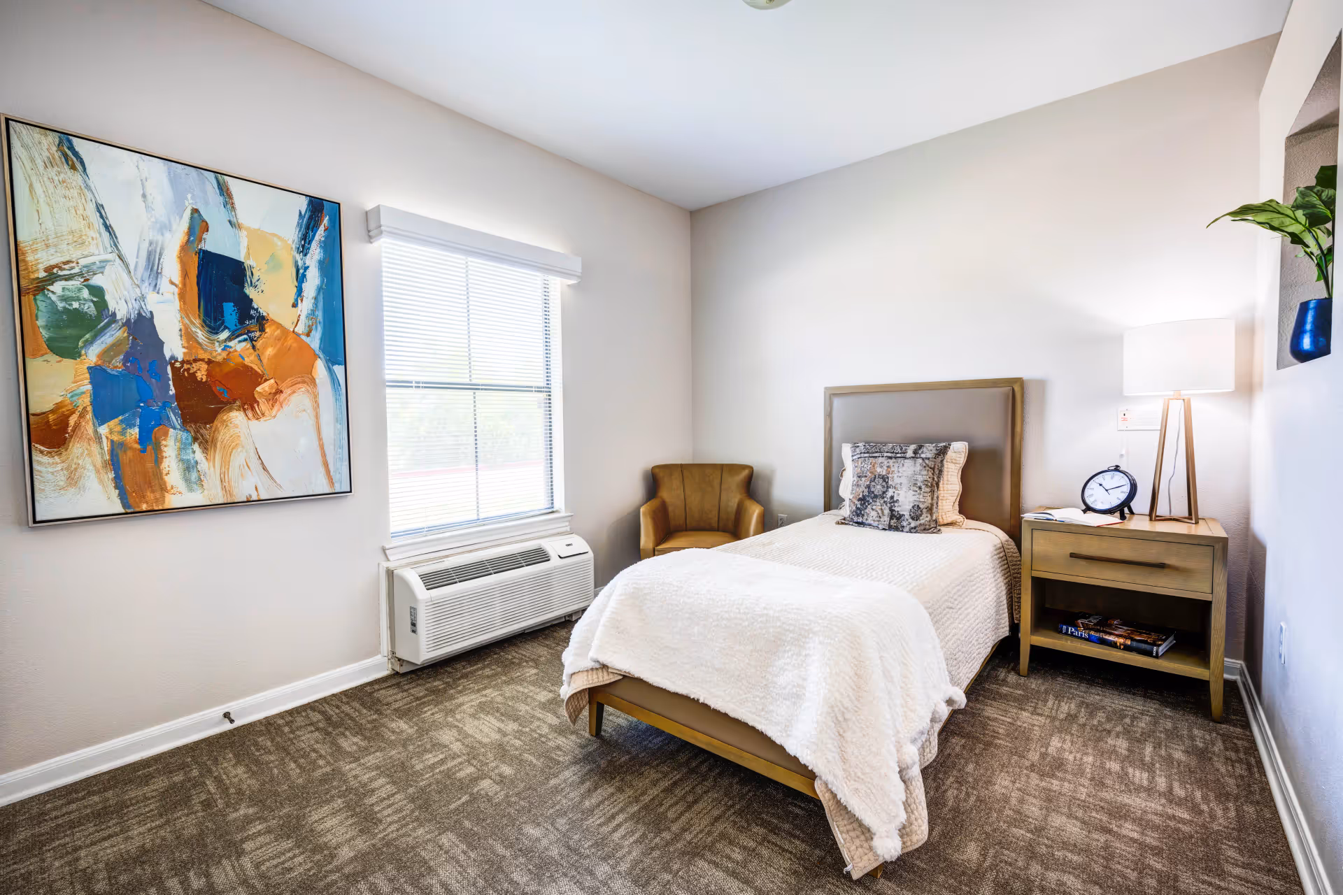 A cozy bedroom with a single bed covered in a white blanket and a decorative pillow. Next to the bed is a wooden nightstand with a lamp, an alarm clock, and a book. A brown armchair is placed in the corner near a window with blinds. There is a colorful abstract painting on the wall and a small plant in a blue pot on a recessed shelf.