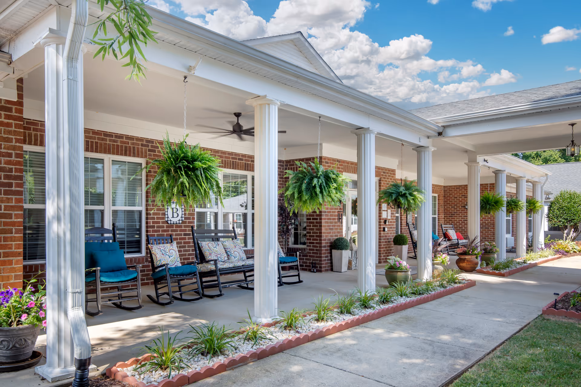Covered porch area of a brick building with white columns, hanging green ferns, rocking chairs with cushions, potted plants, and a clear blue sky with clouds in the background.