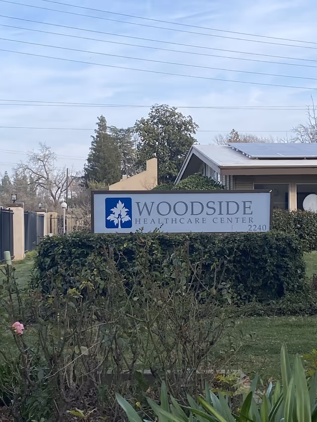 Outdoor view of the Woodside Healthcare Center sign surrounded by greenery and bushes, with a building and trees in the background under a partly cloudy sky.