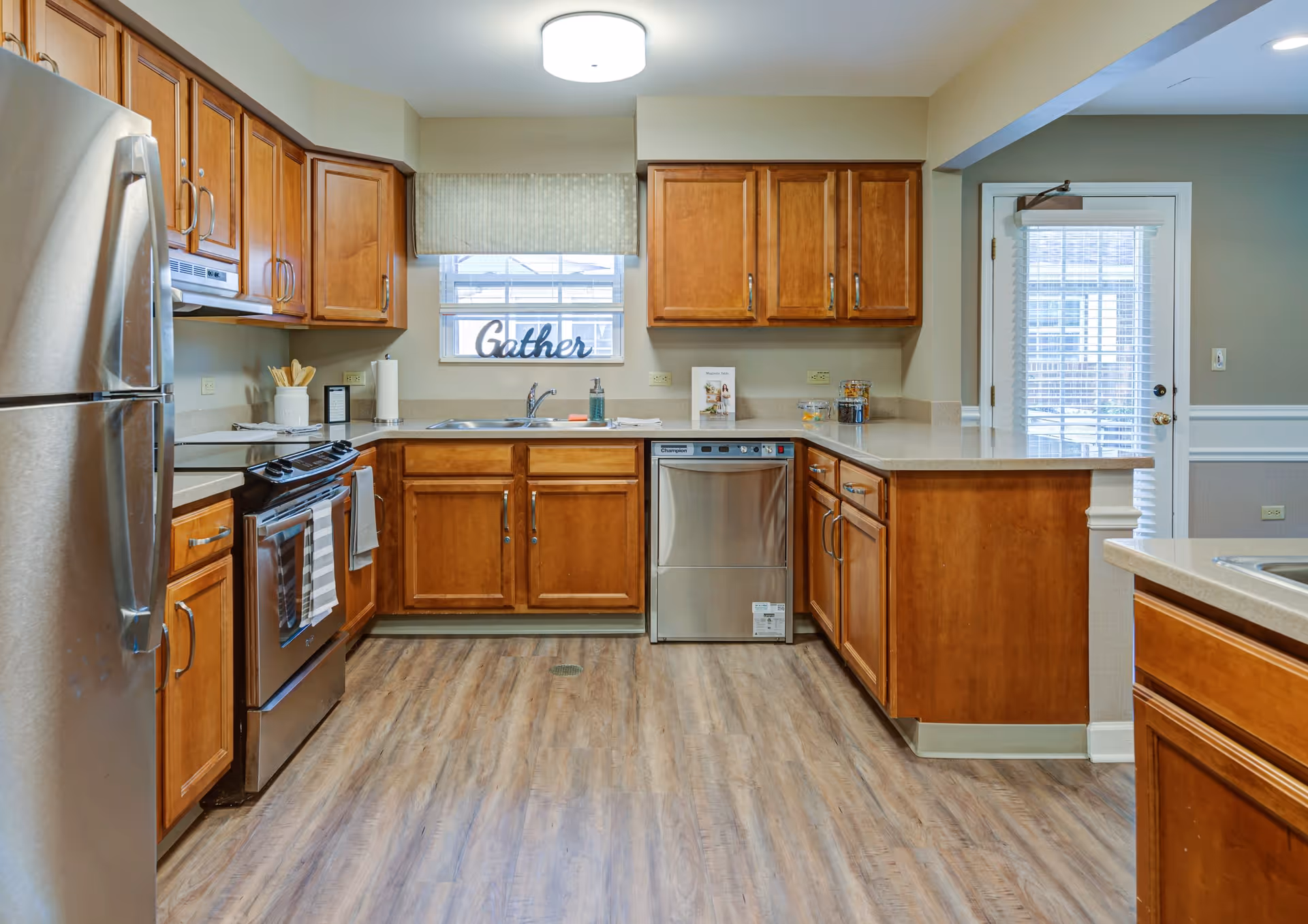 A clean and modern kitchen with wooden cabinets, stainless steel refrigerator, stove, and dishwasher. The kitchen has a light wood floor, a window above the sink with a valance and a decorative 'Gather' sign on the windowsill. There is a door with glass panels and blinds to the right, and the countertops are light-colored with various kitchen items placed on them.