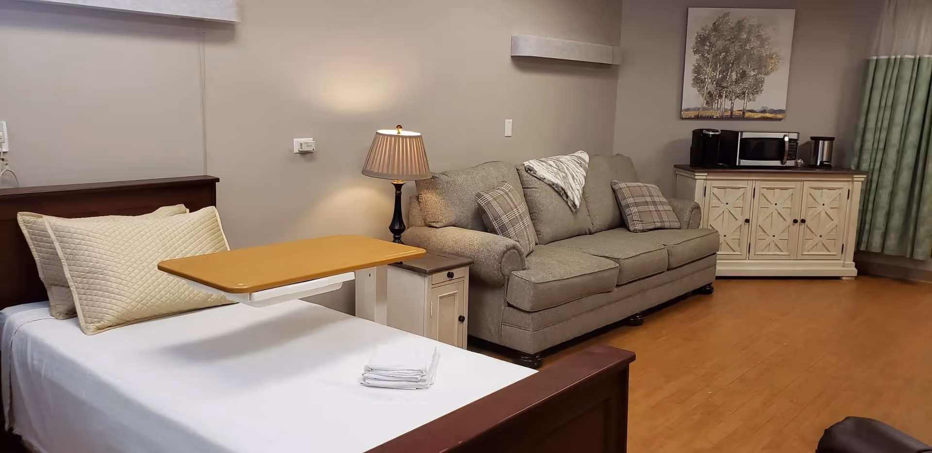 A senior living facility room featuring a single bed with white linens and beige pillows, a wooden overbed table, a beige sofa with patterned cushions, a side table with a lamp, and a cabinet with a microwave, coffee maker, and a painting above it. The room has wooden flooring and green curtains.