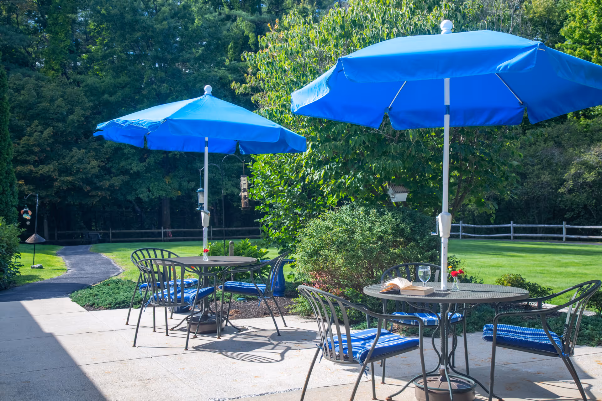 Patio seating area with metal tables, blue umbrellas and chairs overlooking a grassy lawn and trees.